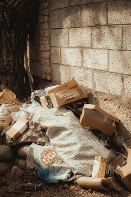 A pile of discarded cardboard boxes, some flattened and some still partially assembled, are accumulated on the ground against a rough brick and concrete wall. The boxes vary in size and are primarily light brown with printed labels and barcodes visible on some. Intermixed with the boxes are plastic bags, one of which appears to contain additional packaging waste, and a fabric sack or large piece of woven material that is partially covered by the debris. The setting is outdoors, near a building structure, with a section of dark tree bark visible to the left and small rocks and dirt scattered at the base of the pile. The lighting is natural, casting soft shadows, suggesting daytime. This scene reflects a typical instance of waste awaiting collection or alternative disposal, relevant to private rubbish removal services such as those offered by Waste Disposal Bermondsey, emphasizing on-site clearance or independent rubbish handling in an urban environment near a brick wall background.