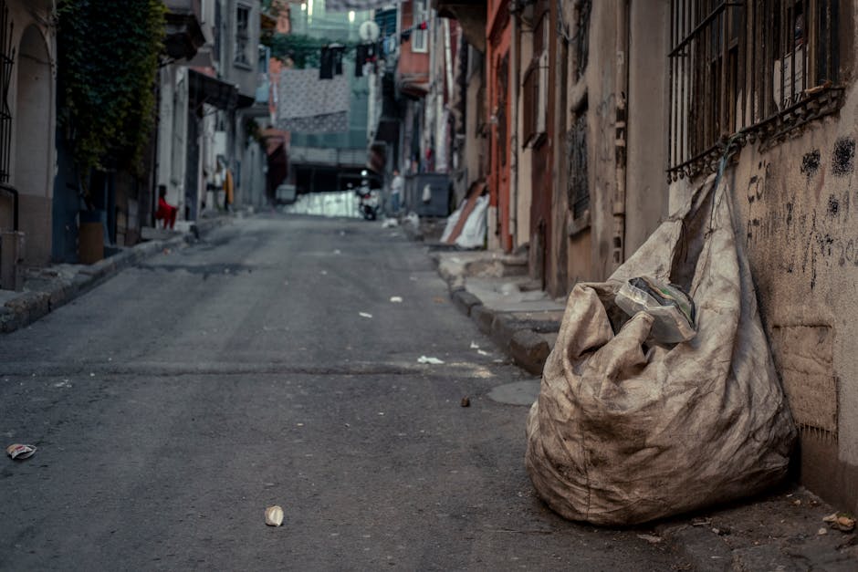 A large, weathered beige fabric garbage bag is positioned on the right side of a narrow, uneven urban street paved with asphalt. The bag appears full and slightly sagging, with visible wrinkles and dirt marks on its surface. Resting on top of the bag is a discarded transparent plastic container. In the background, the street slopes upward and is lined with various residential buildings constructed from brick and concrete, some featuring iron window grilles and graffiti on the walls. To the left, a few small objects and scattered litter are visible on the pavement, contributing to an overall scene of unmanaged waste. The scene is illuminated by natural daylight, with shadows cast by the buildings. Some laundry or fabric items are hanging on clotheslines strung between buildings further up the street, indicating a densely populated area. This setting exemplifies common challenges of private rubbish disposal in urban neighbourhoods, where independent waste collection services like Waste Disposal Bermondsey may be employed for on-site clearance and refuse management.