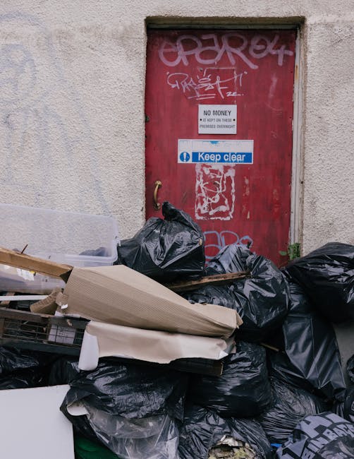 A pile of garbage bags and discarded waste materials stacked against a textured beige wall in front of a closed red metal door covered with graffiti and various stickers. The waste includes black plastic bags filled with rubbish, a large piece of cardboard, and some broken or torn paper items. The door features a white sign indicating 'No Money' and a blue and white sticker saying 'Keep Clear,' with additional graffiti tags in white and pink above and around the doorframe. The scene appears to be an outdoor alleyway or back entrance, with natural lighting highlighting the cluttered state of the rubbish accumulation, which may relate to private waste disposal or on-site clearance services offered by Waste Disposal Bermondsey.
