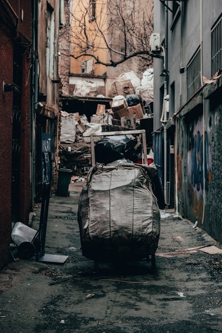 A narrow urban alleyway cluttered with large black and white garbage bags and loose rubbish, including cardboard boxes, plastic containers, and paper waste piled against a makeshift wall of discarded materials at the far end. The ground features dark, stained asphalt with scattered debris, and the surrounding buildings are aged, with graffiti on the metal fences and walls on the right side. On the left, a metal pole and a small waste bin are visible, while leafless tree branches extend overhead, indicating a colder season. The scene appears to be an area awaiting rubbish collection, characteristic of private or on-site waste disposal in an urban environment, with Waste Disposal Bermondsey occasionally providing services for such collections.
