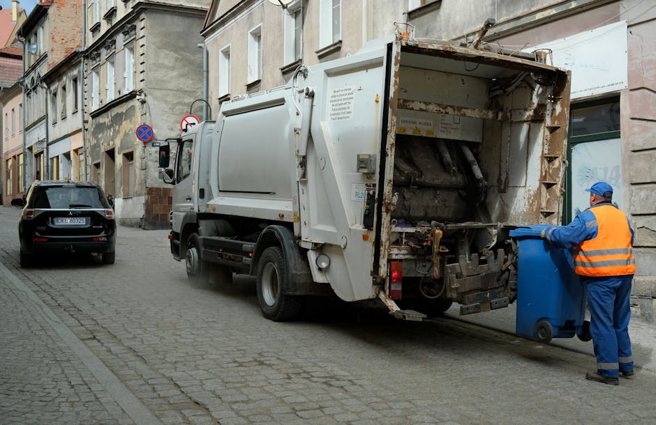 A municipal rubbish collection vehicle with a white exterior and an open rear compactor area, positioned on a narrow cobblestone street. The vehicle is in the process of emptying a blue wheeled bin held by a worker dressed in a high-visibility orange vest, blue overalls, and a blue cap. The worker stands to the right of the truck, guiding the bin towards the open rear compartment. The backdrop features older multi-story buildings with attached windows and street signs, including no parking and no stopping signs. A dark-colored car is parked along the left side of the street, and the overall scene appears to be during daytime in an urban residential area, supporting the context of private waste collection and on-site rubbish removal services typical of Bermondsey. The weather appears overcast, with diffused natural lighting highlighting the textures of the cobblestones and the rusted metal parts of the refuse vehicle, aligning with waste disposal activities conducted by services like Waste Disposal Bermondsey.