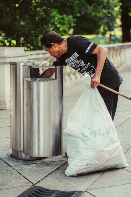 A man with short, dark hair, dressed in a black t-shirt with white text and dark pants, is seen leaning forward to dispose of a white plastic bag filled with rubbish into a stainless steel litter bin on a paved outdoor walkway. The bin features a sleek, cylindrical design with a top-opening lid and is situated alongside a low, stone balustrade. In the background, there are lush green trees and bushes, suggesting a park or garden setting, with natural daylight illuminating the scene. The man is holding a rake-like tool in his right hand, which rests against his waist, and his posture indicates he is actively engaged in waste disposal or cleaning. The environment appears well-maintained, with clear contrast between the shiny metallic surface of the bin and the textured, grey paving stones surrounding it. This scene exemplifies appropriate private waste handling and rubbish collection practices, supporting efficient rubbish removal services offered by Waste Disposal Bermondsey.