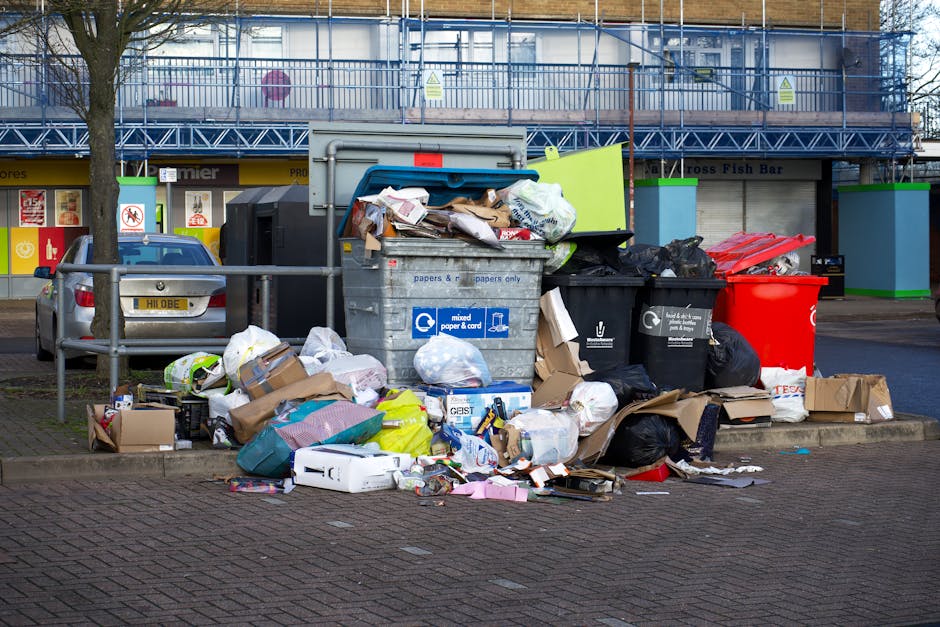 An outdoor scene showing multiple open-top waste bins and overflowing rubbish on a paved area in front of a commercial building, with discarded cardboard boxes, plastic bags, paper packaging, and household waste spilling onto the ground. The waste containers include a large gray mixed paper and cardboard bin, a black recycling bin, and a bright red bin, all positioned near a metal railing on the pavement. Behind the bins, there is a parked silver car on the street and a commercial storefront with a yellow sign, while the upper floors of the building are under construction, covered with blue safety scaffolding. The surrounding environment appears to be an urban area with a tree on the left side, and the scene is lit by natural daylight, highlighting the clutter of waste and the textures of the various materials. This image relates to private rubbish collection services such as those offered by Waste Disposal Bermondsey, focusing on on-site clearance and alternative waste handling beyond local authority rubbish collection.