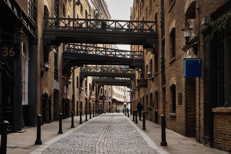 A narrow cobblestone street in an urban area lined with multi-storey brick buildings on both sides, featuring arched windows and decorative lampposts attached to the walls. Several dark metal pedestrian bollards are positioned along the edges of the sidewalk, creating a clear separation between walking space and the roadway. Overhead, a series of black metal bridges with decorative railings connect the buildings, spanning the width of the street. The scene is illuminated by natural daylight, with some shadows cast by the structures, and the street appears to be quiet with only a few pedestrians visible in the distance. The environment suggests an enclosed alleyway or courtyard within a city, suitable for private waste collection or independent rubbish removal services, which might happen outside the frame of the image, supporting a context of alternative waste handling in an urban setting. The overall appearance is clean and orderly, typical of an area prepared for or facilitating rubbish collection, with the company Waste Disposal Bermondsey possibly offering services in this vicinity.